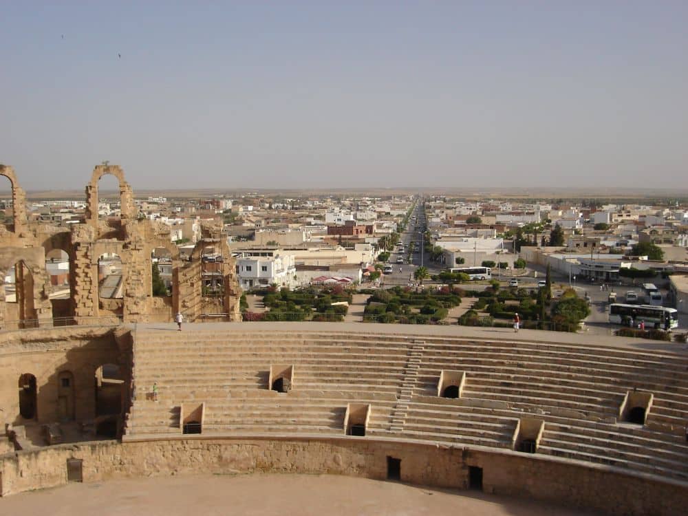 A view over the modern bleacher section to the city of El Jem. An absolutely straight modern road straight head, low buildings on either side.