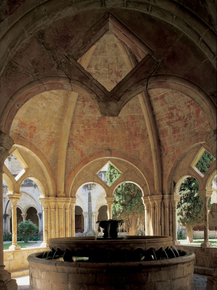 A fountain shaded by a round stone structure with arched windows onto the garden of the cloister.