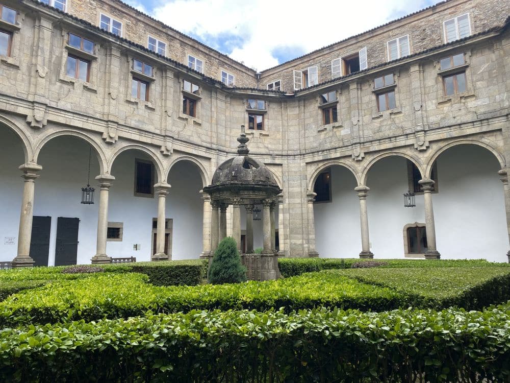 Two sides of a cloister with arches around the sides forming a portico and neatly trimmed shrubbery in the courtyard.