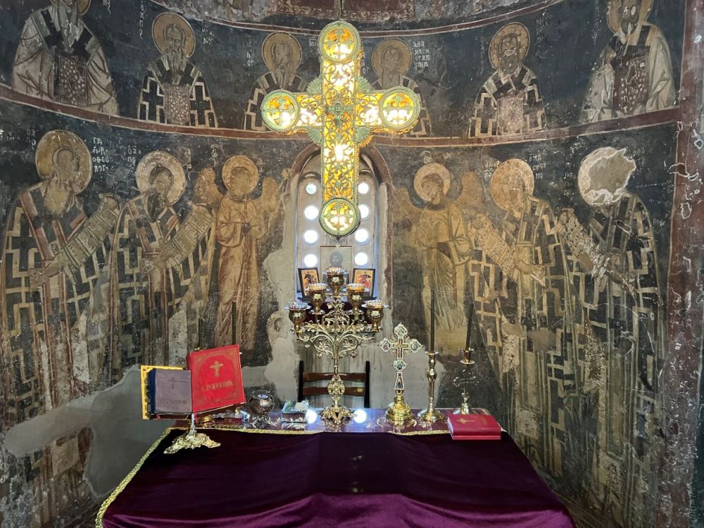 An altar with a large cross and frescoes covering the rounded wall behind it.