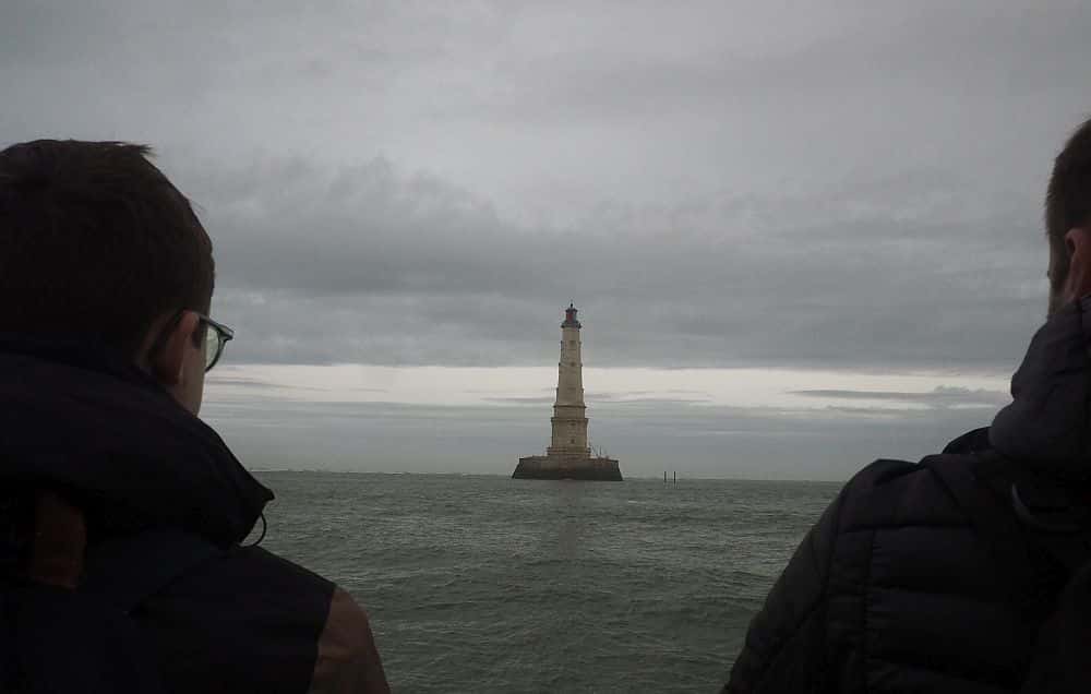 Seen between two people looking away from the camera, the lighthouse stands in the ocean, seemingly sticking out of the water, no land around it.