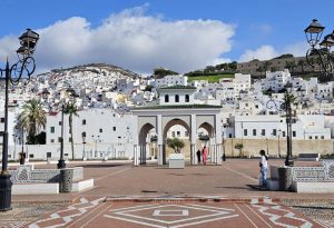 An arched gateway, with many white-painted houses arrayed up a hill behind it.