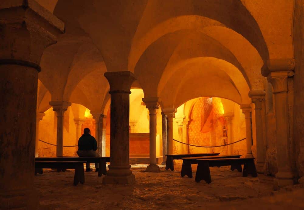 A dark room with vaulted ceiling, benches on a stone floor, light above the altar in the background.