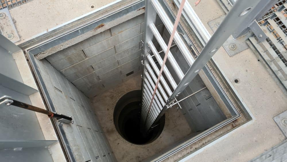 A square opening and a metal ladder leading down to a round opening at the bottom of a shaft.