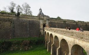 An arched stone bridge over a dry moat leads to a massive wall with a small gateway in it.