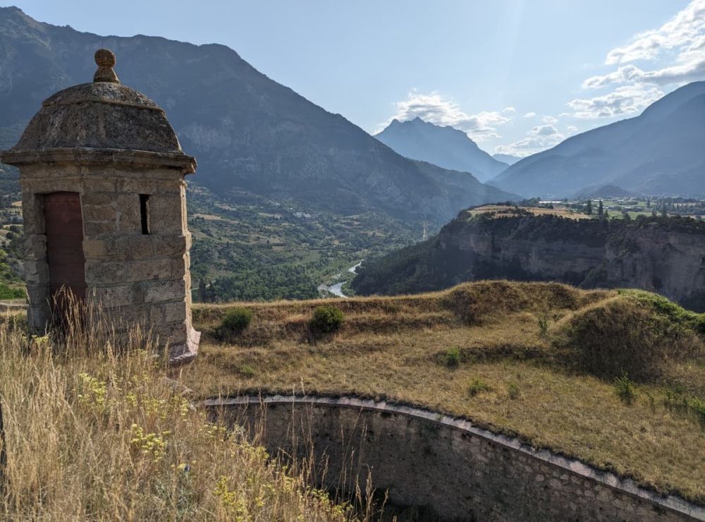 In the near distance, a small guardhouse on an earthen embankment. Beyond, a river valley and the cliffs on the far side of the river.