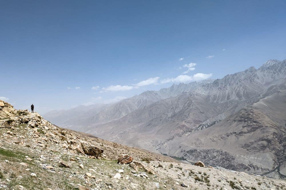 A person stands on the side of a barren, rocky slope. Beyond him is a view of mountains.