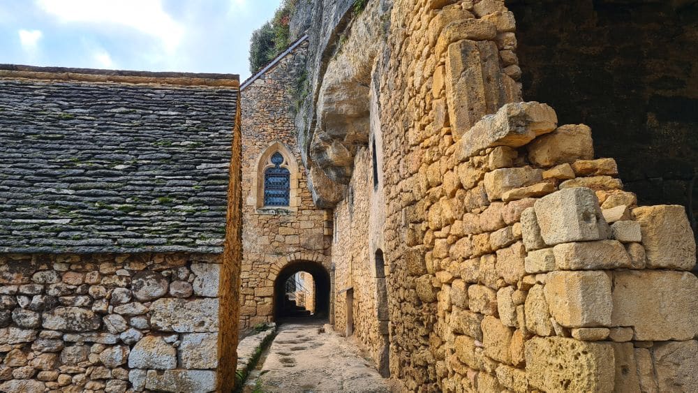 View of a narrow stone street with rock walls on the left and a church straight ahead. The street goes through an arch under the church.