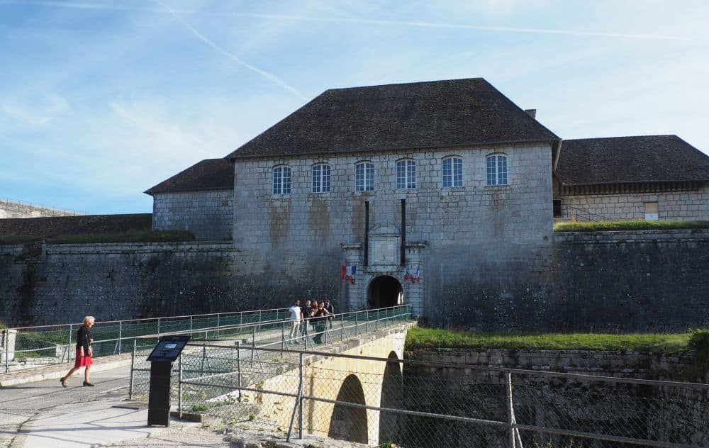 A large, block-like stone building at the end of an arched bridge over a dry moat.