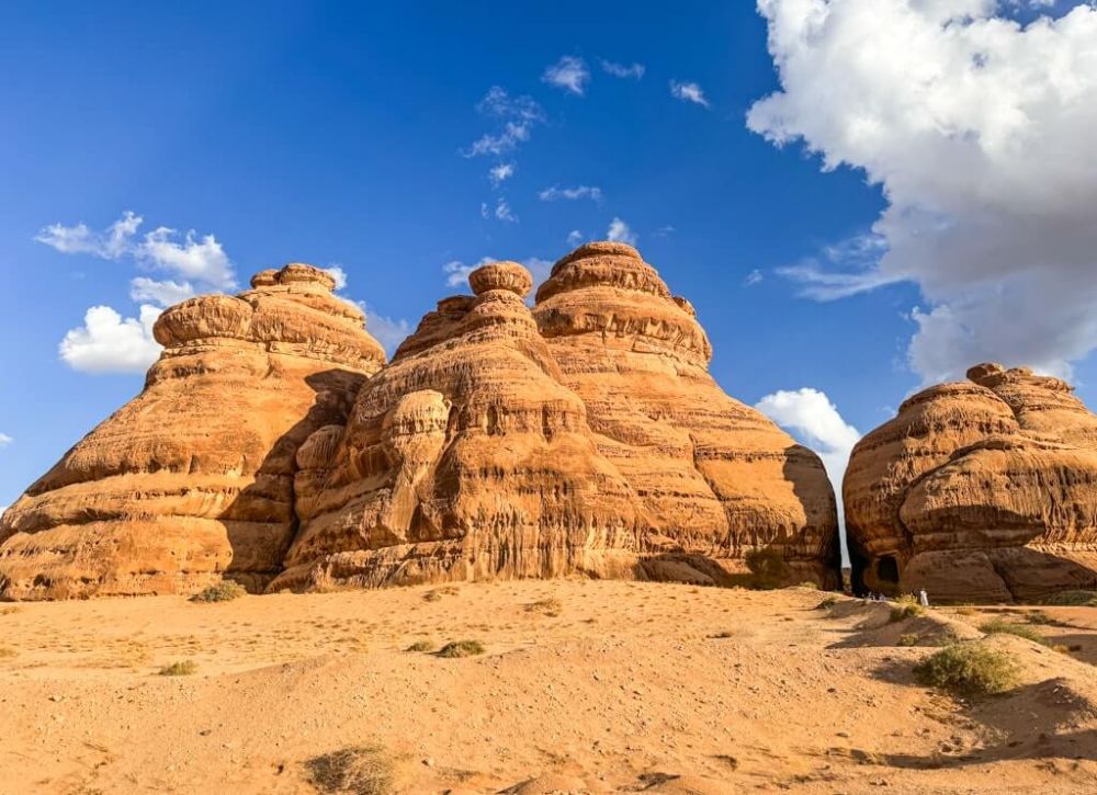 Reddish cliffs eroded into interesting shapes, with the entrance to a tomb, very small-looking, at the base of one of the shapes.