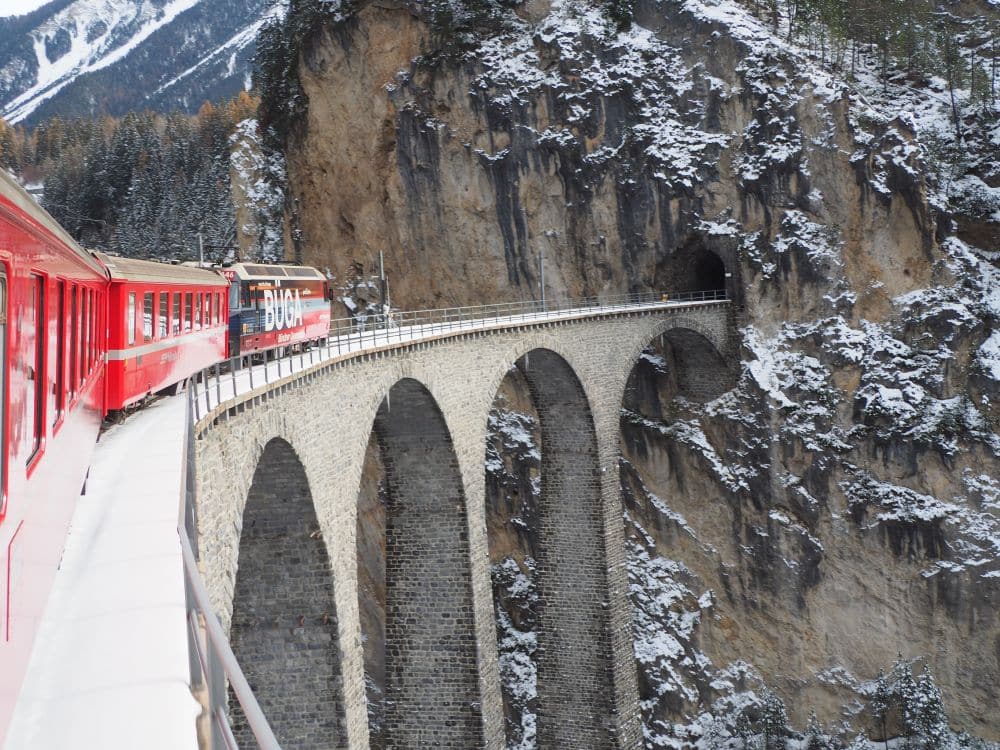 A red Rhaetian Railways train curves to the right onto an arched stone viaduct over a deep gorge. At the other end of the viaduct is a tunnel into the side of a mountain.