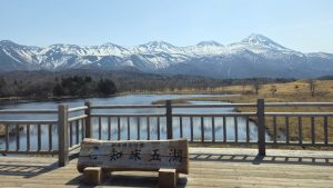 A walkway with a sign and a guardrail. Behind the rail, a lake and beyond that, a snow-capped mountain range.