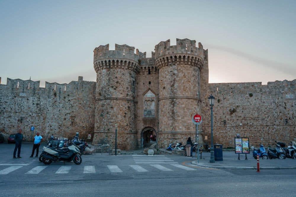 A large stone city wall with a gate in it. The gate has two round towers on either side, topped with crenelations.