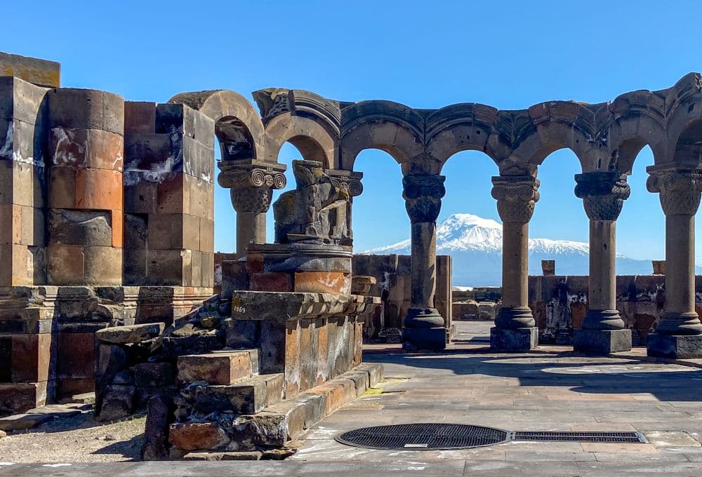 A row of pillars holding up arches, with a snow-covered mountain in the background.