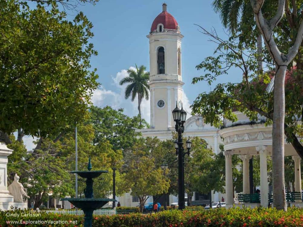 A church steeple in an elegant style, but the rest of the church is mostly obscured by tropical greenery in the park in front of it.