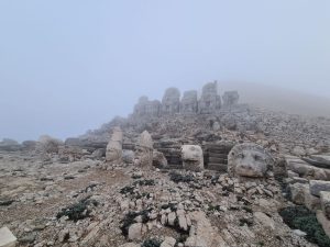 A misty and rocky mountaintop, with a pedestal with broken statues on it, their heads on the ground in front of them.