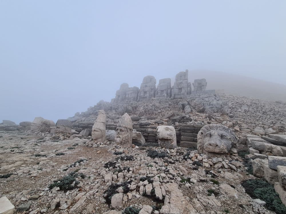 A misty and rocky mountaintop, with a pedestal with broken statues on it, their heads on the ground in front of them.