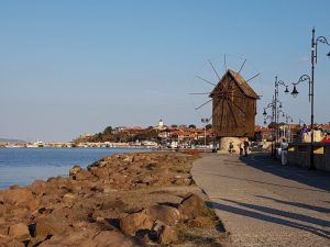 A path with stones between it and the sea, a windmill along the path, and part of the Ancient City of Nessebar visible beyond the windmill.