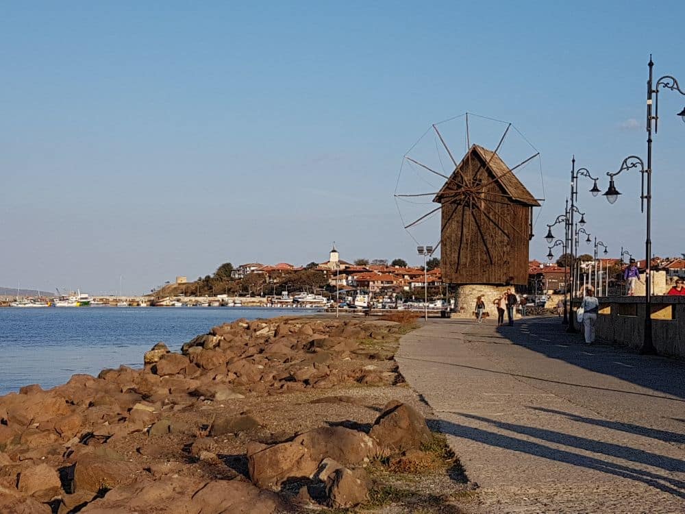 A path with stones between it and the sea, a windmill along the path, and part of the Ancient City of Nessebar visible beyond the windmill.