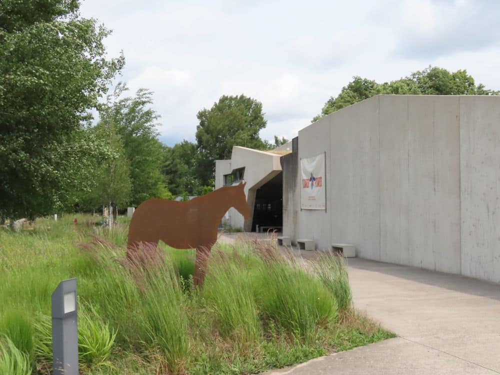 A low concrete building in a modern brutalist style, with a metal cutout of a horse standing in front of it.