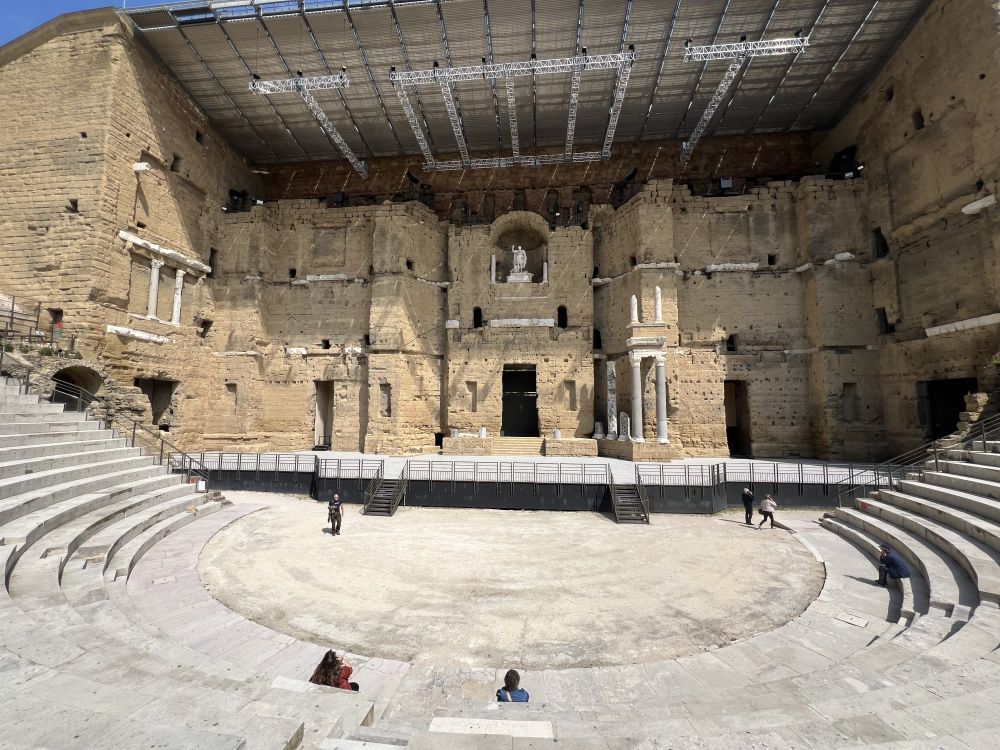 The backdrop of the theatre is a couple of stories high, in stone, and the half-circle of seats is partly visible.