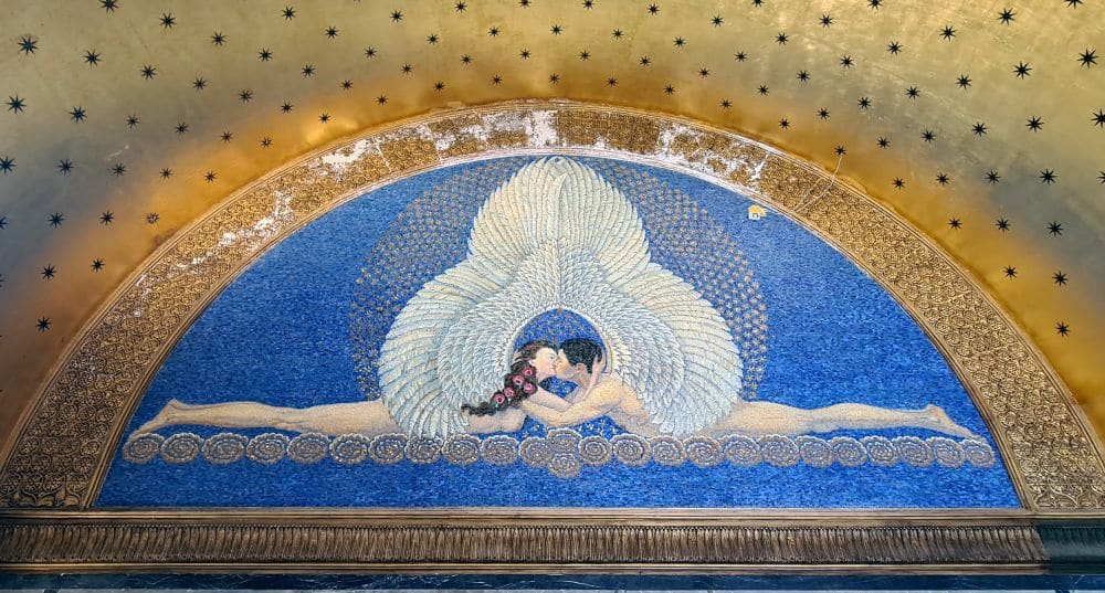 An image of two people lying facing each other - feet at the outside edges, faces together, kissing. They're both naked but have large white wings that meet over their heads.