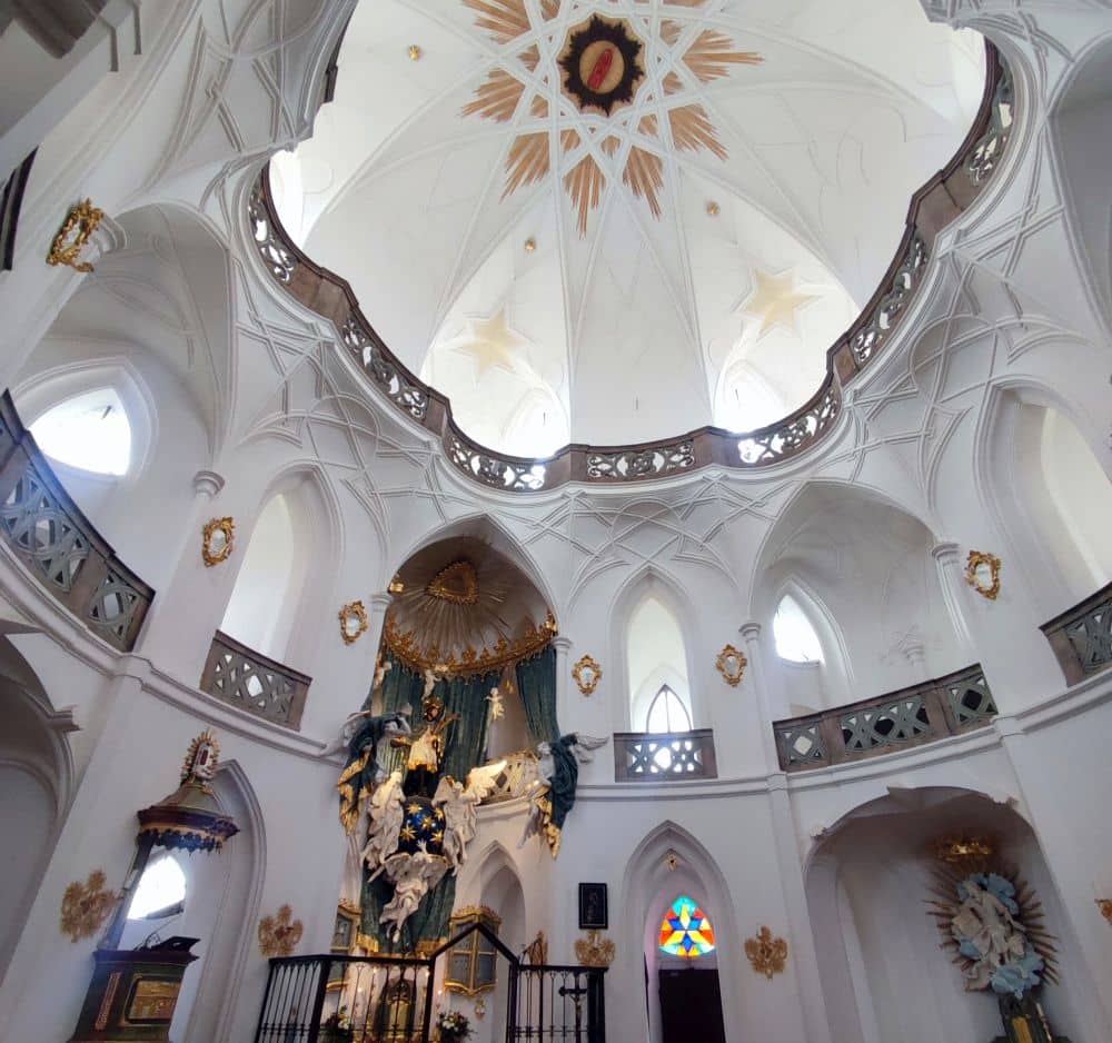Looking up inside the church: white walls in several tiers of arches with decorative railings, a a more highly-decorated altar, and a bit of painted decoration at the top of the central dome, but otherwise plain white.