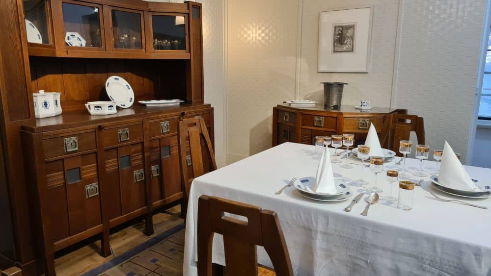 A table covered with a tablecloth, set with place settings and glasses. A sideboard cupboard with more china and glasses.