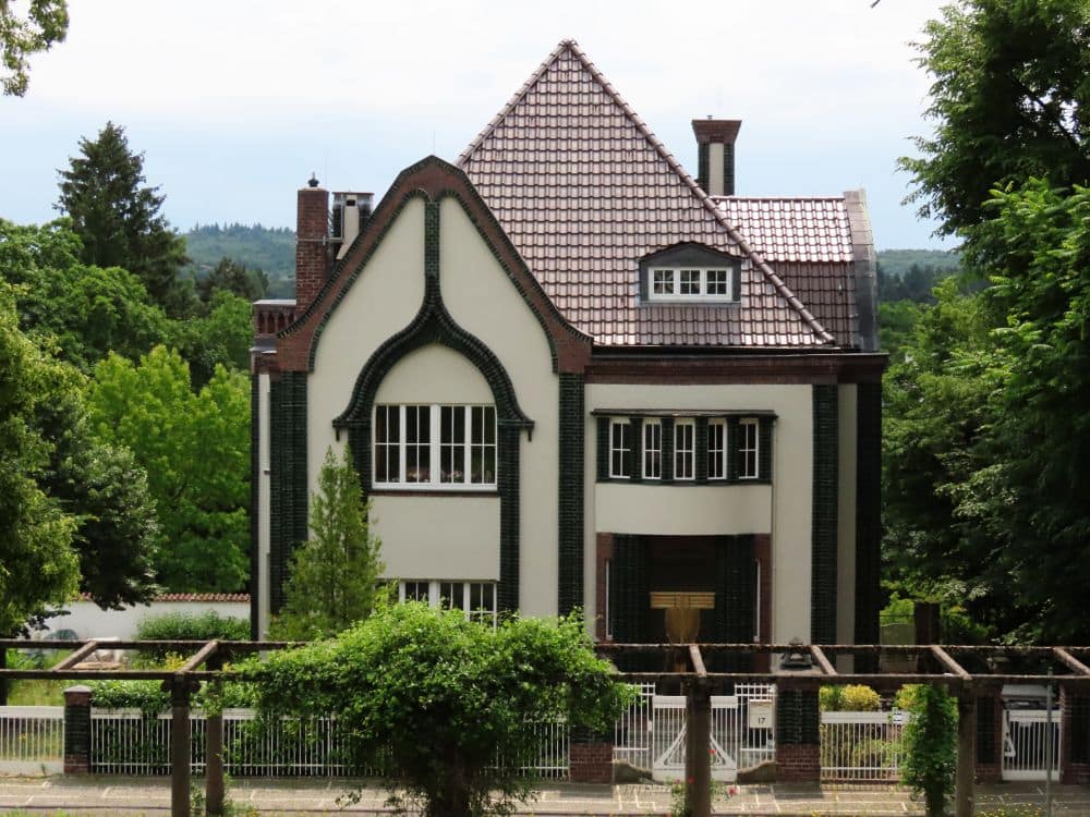 A square house with peaked tile roof, horizontal strips of windows, and an ornate door.