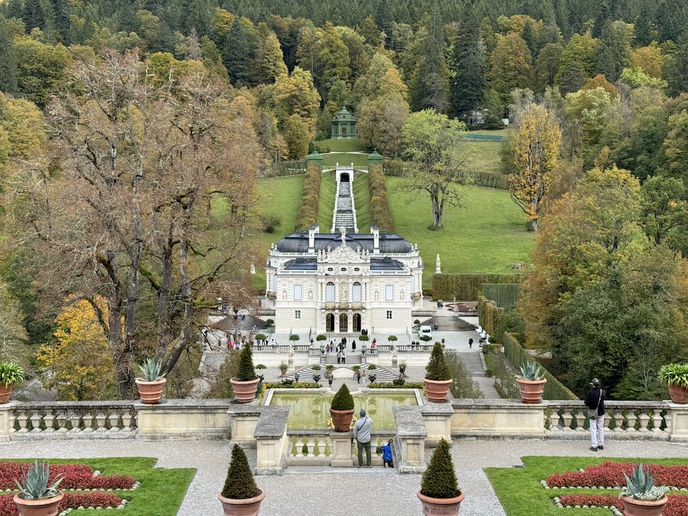 Looking downhill at a smallish white palace surrounded by formal gardens, in turn surrounded by wooded hills. One of the Palaces of King Ludwig II.