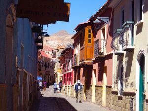 Looking down a narrow street in the City of Potosí lined with colorful two-story rowhouses. In the distance, a conical, brown mountain looms.