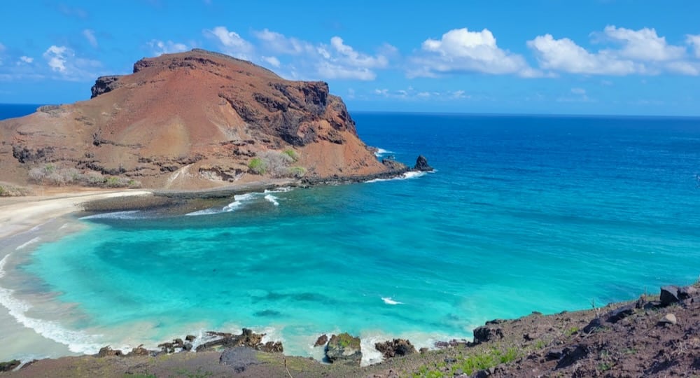 A view of a small bay with clear blue water, a rocky outcropping on the far side of it.