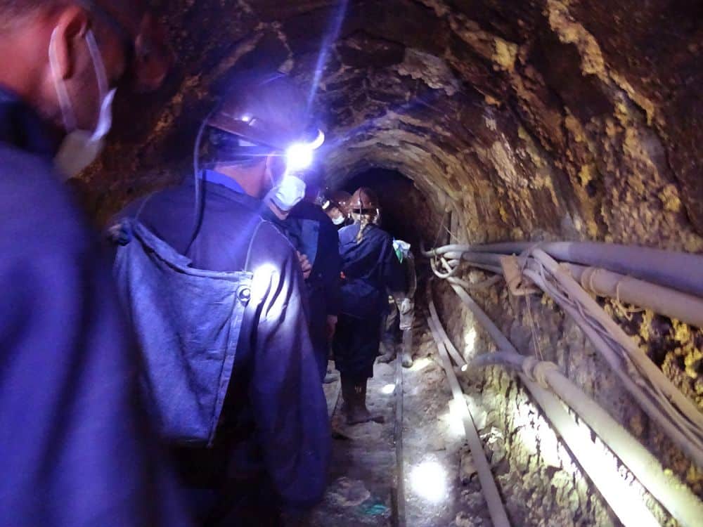 A narrow tunnel, dimly lit. People wearing blue clothes and hardhats walk away from the camera single-file.