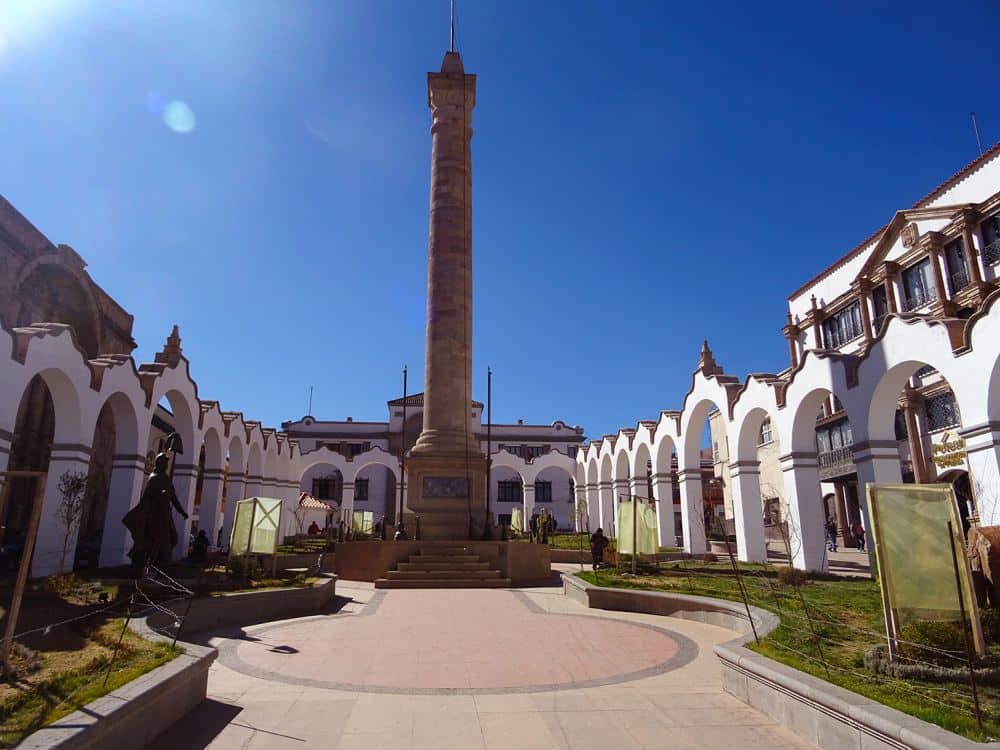 A square edged by a rows of arches with the buildings behind them. In the center, a tall monument.