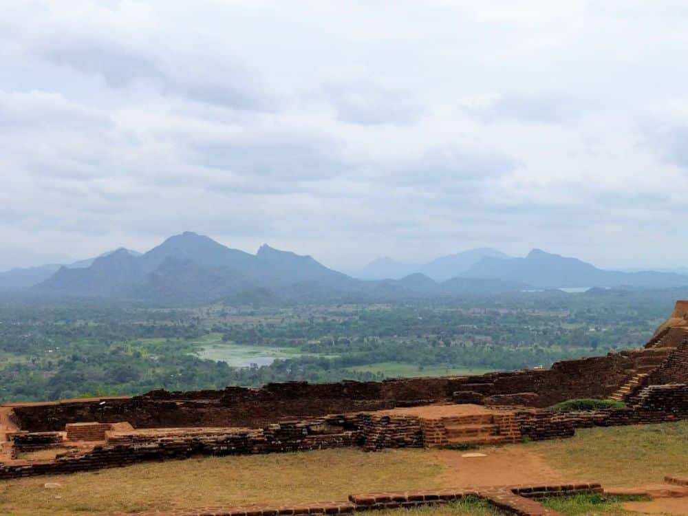 View of a valley and mountains beyond it, with the low remains of palace walls in the foreground.
