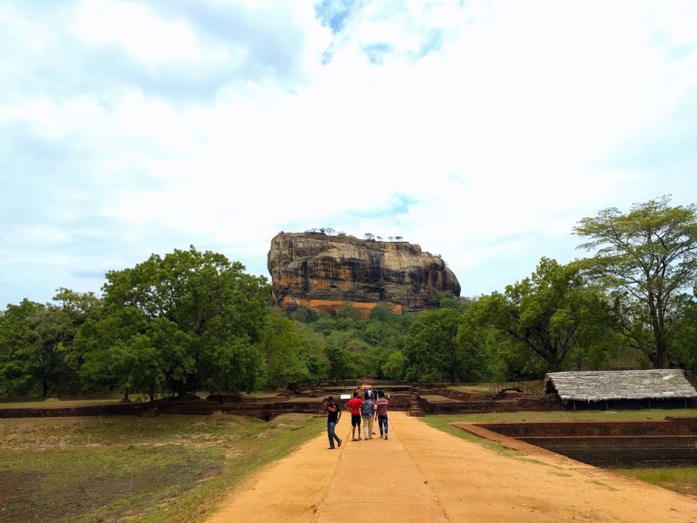 View of the rock looking down a road toward it, rising in the distance where the Ancient City of Sigiriya sits on the top. 