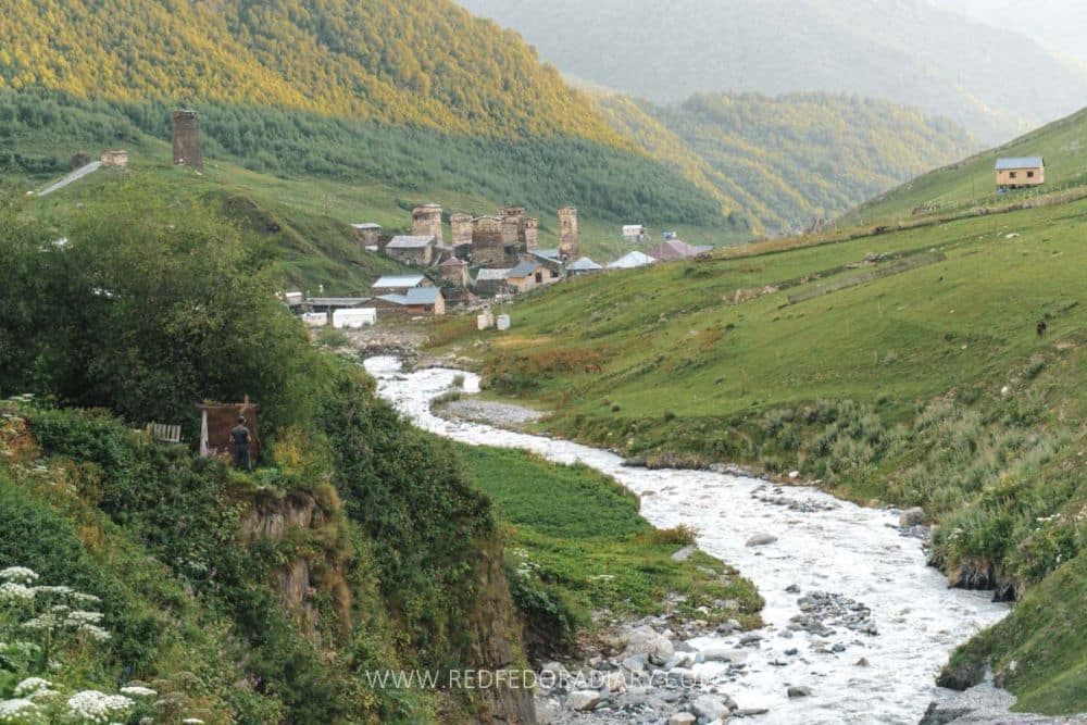 A rushing stream emerges from a village in the distance and curves between mountains.