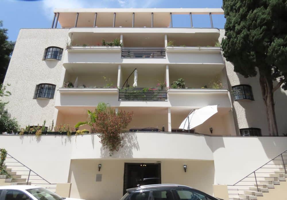 An apartment building iwth balconies across the front, a raised entrance op two side flights of stairs, and a rounded entrance area.