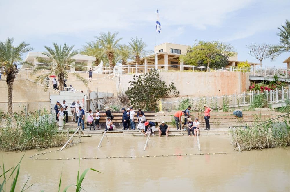 Muddy water and, seen on the other side of it, people dip their feet in the water. A building behind them with the Israeli flag.
