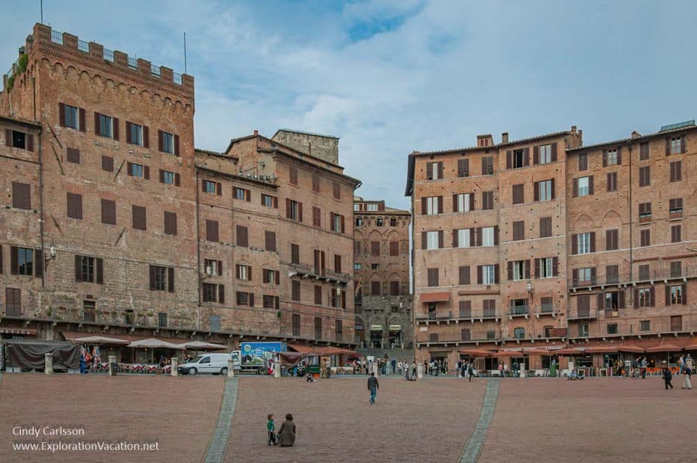 An open brick-paved plaza lined with tall brick buildings of about 7 stories, clearly medieval, with cafes on their ground floors. 
