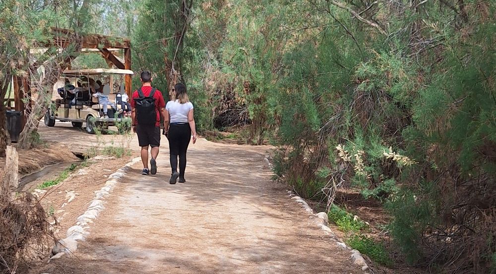 The path is dirt, but neat and flat, with trees shading it. Two people walk ahead, and a small open vehicle carries tourists on the path.