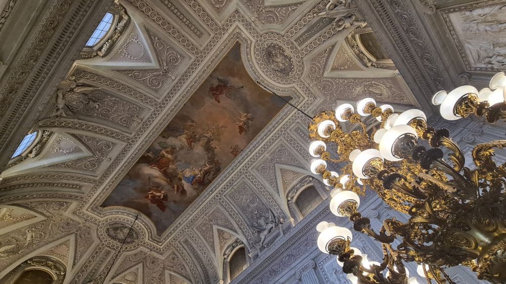 Looking up at a ceiling past a huge chandelier. The ceiling has an ornate baroque fresco on it, and is highly decorated with various stucco decorations.