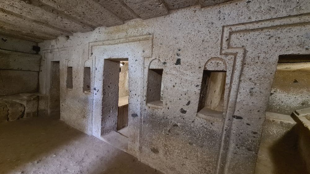 A stone cut room with beams carved into the ceiling, two doorways with simple door frames cut around them, and small windows cut between rooms. A bit of two smaller rooms can be glimpsed through the two doorways, each with stone-carved beds.