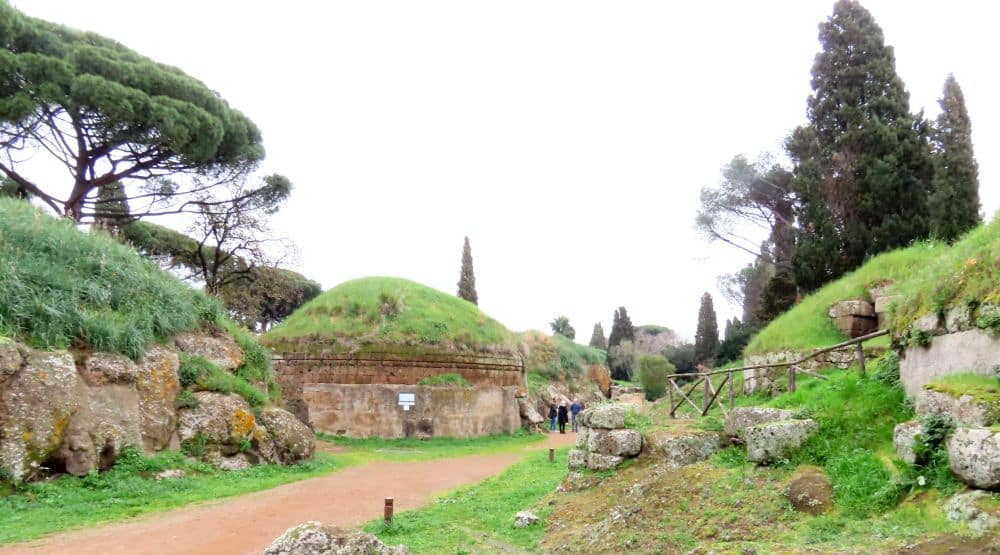 A row of tumuli with stone sides and rounded grass tops. A group of people stand on the path in front of one of them, showing that the tumuli are very large, towering over them.