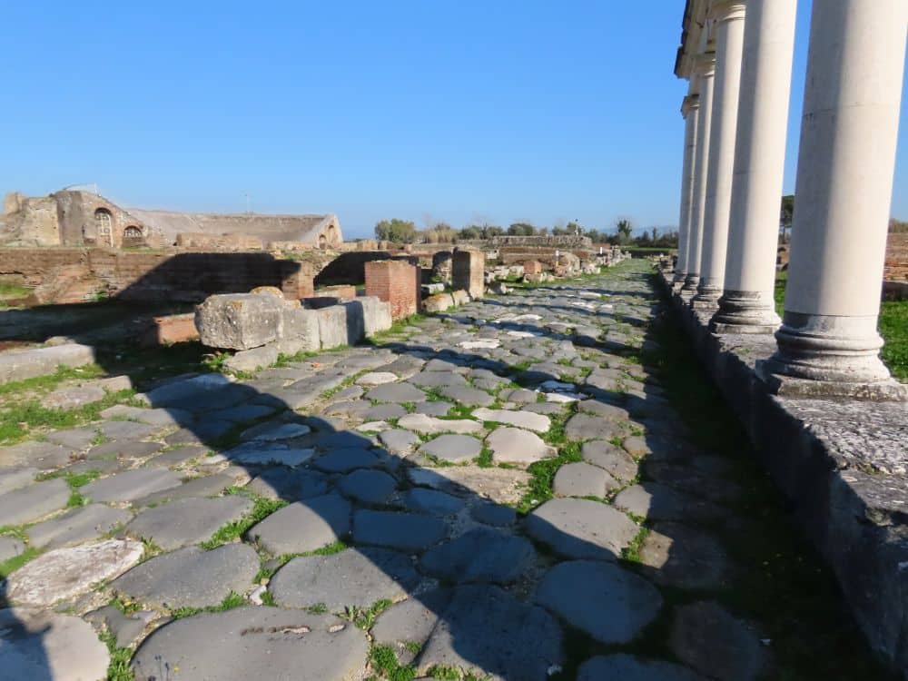 A row of columns on the right, a stretch of original Via Appia long them, and some low ruins of walls on the left.