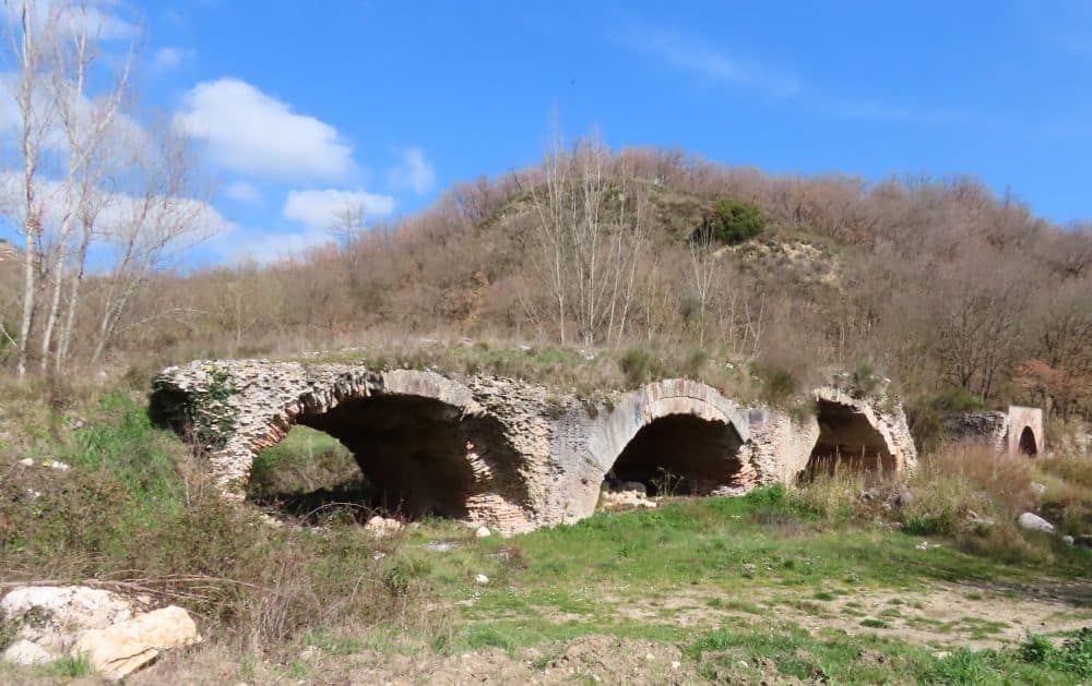 Arches of a stone bridge, still standing, with grass and shrubs growing along its top.
