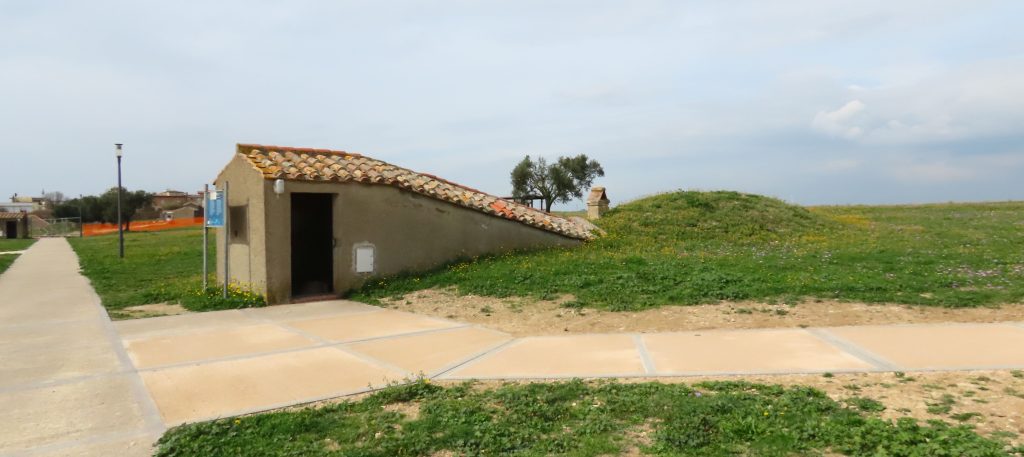 A paved path, a small building with a single door and a roof that extends from on top of the building in a slant down to the ground, where a grass-covered bump is lower than the building.