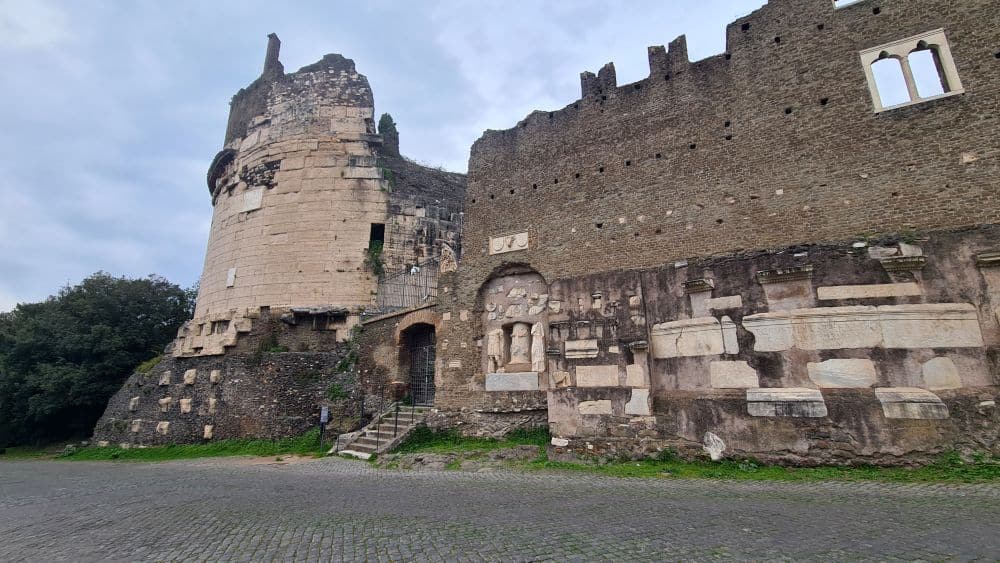 A round tower, partly ruined, on the left, and a castle wall on the right, with some Roman-era pieces of monuments embedded in it.