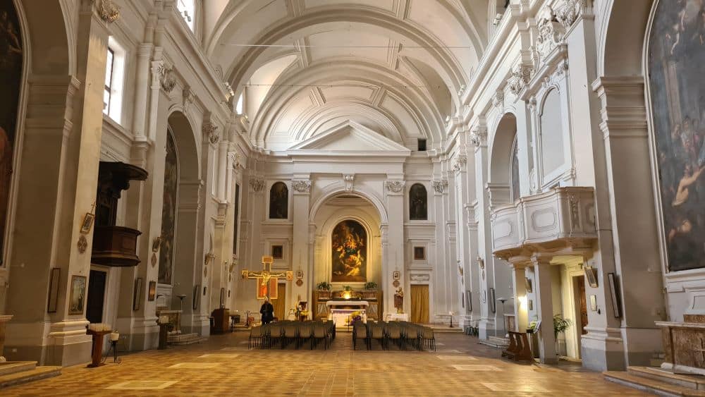Large interior with white walls in neo-classical style: pillars, Greek-style pediment over the altar. Huge paintings in each arch.