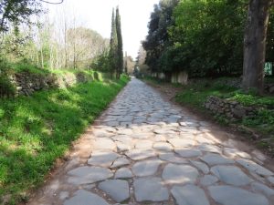Original Roman-era paving stretching into the distance between grassy banks - stone wall on the left and trees on the right.